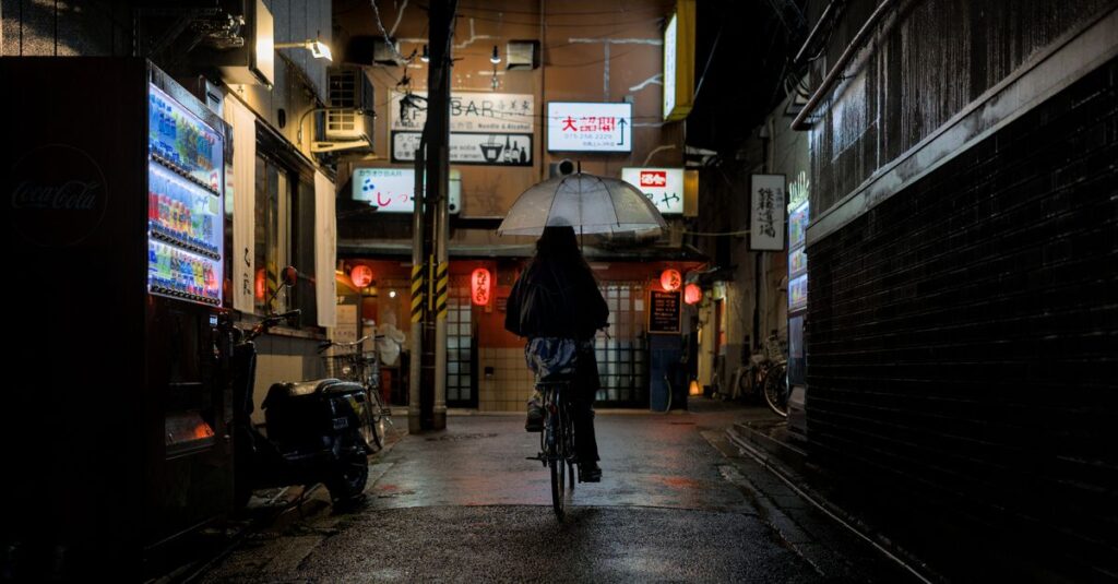 夜間に傘を差して片手運転をする自転車のイラスト。無灯火や傘さし運転の危険性を表現。